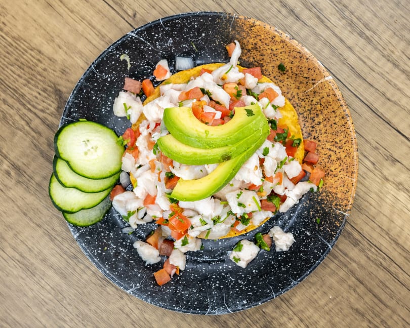 Ceviche tostada with avocado slices, diced tomatoes, and cucumbers on a speckled plate.