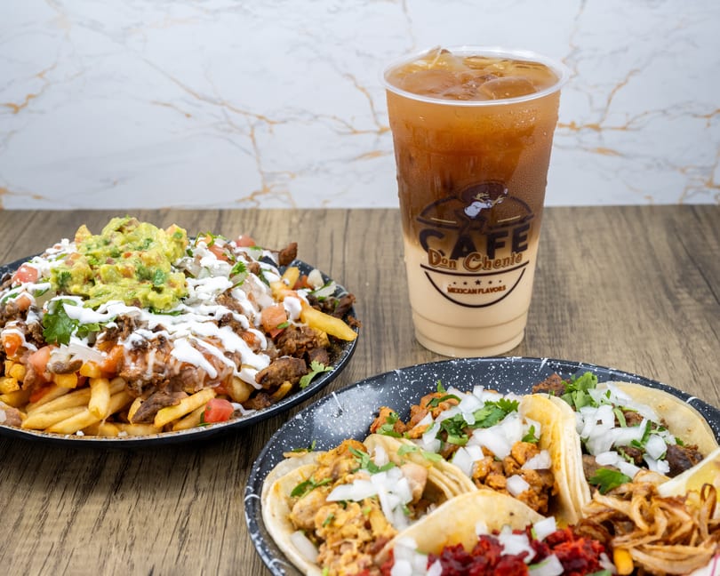 Plates of loaded carne asada fries, assorted tacos, and an iced coffee drink on a wooden table.