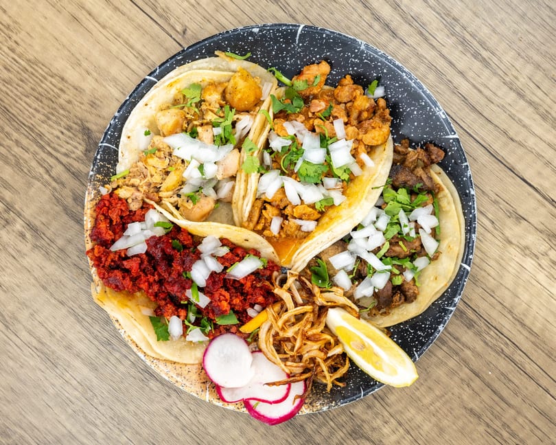 Four tacos with various fillings, onions, cilantro, fried onions, radishes, and a lemon wedge.