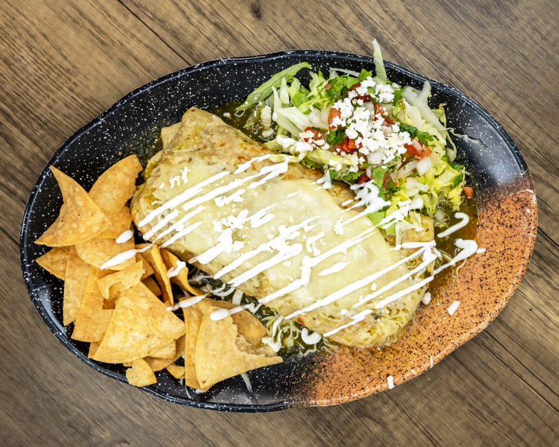 Two cheesy burritos covered in green sauce, with a side of tortilla chips and fresh salad.