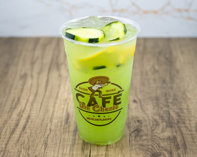 A Cafe Don Chente Mexican Flavors drink, light green with cucumber slices and ice, on a wooden table.