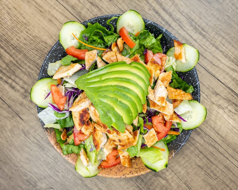 Overhead view of a fresh salad with grilled chicken, sliced avocado, tomatoes, and cucumbers.