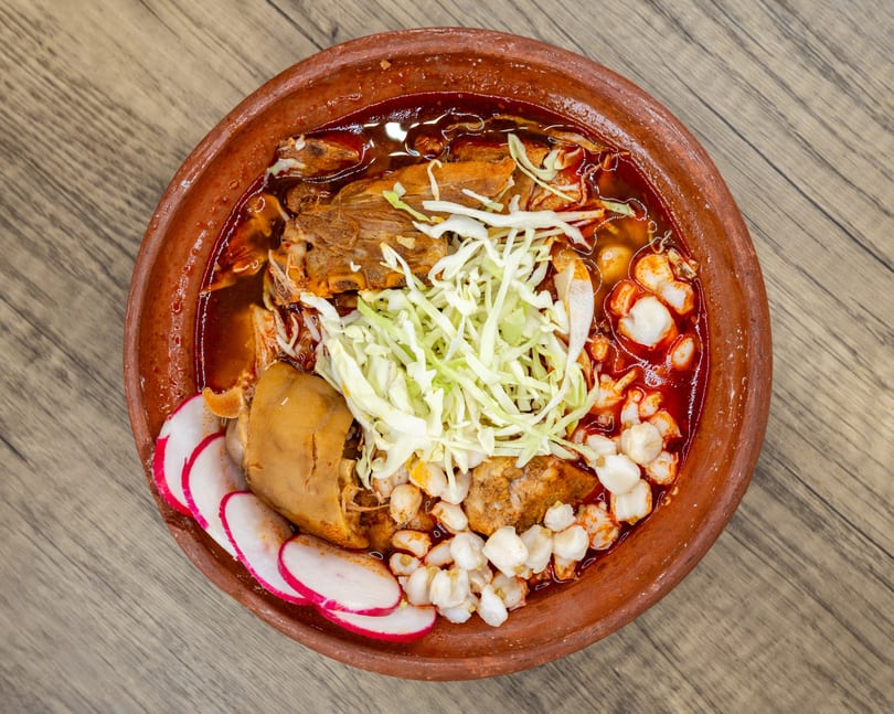 Overhead view of a pozole rojo in a clay bowl, garnished with cabbage, radishes, and hominy.